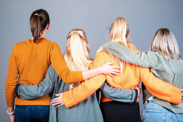 Vier junge Frauen stehen in einer Reihe und halten sich fest. Man sieht sie von hinten. 